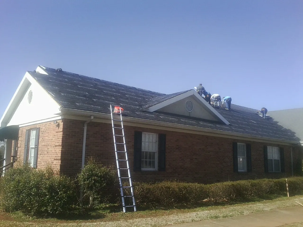 Crew tearing shingles off a brick ranch roof with a ladder set — Emergency Roof Repair work by our crew in Edgerton